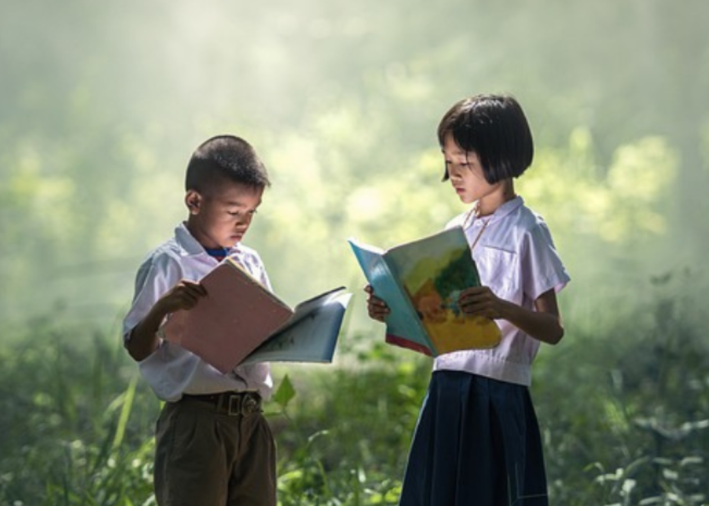 Two school children reading books in a beautiful garden while standing up.